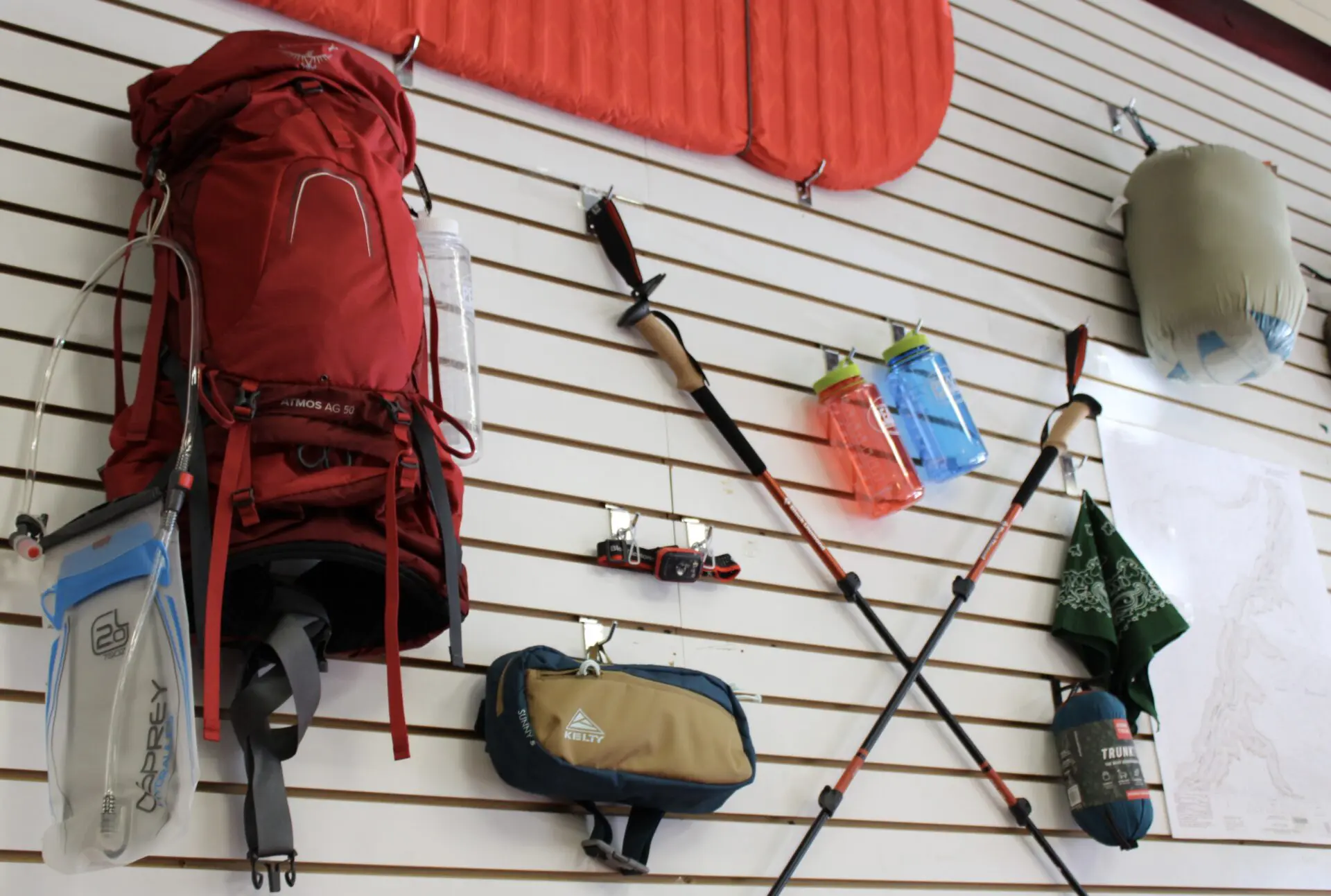 Various camping and hiking gear, including a red backpack and hydration packs, displayed on a white wall with a map.