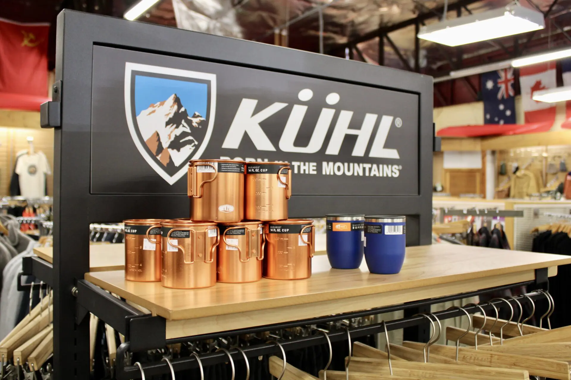 Copper-colored metal tumblers on display in front of a kühl retail store sign with clothing racks and flags in the background.