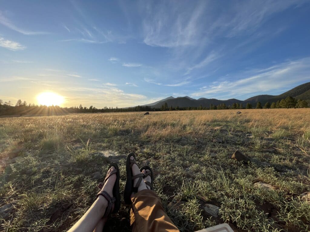 A person’s feet on the grassy field near the mountain while the sun shines bright