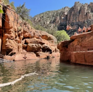 People cliff jumping into a river surrounded by tall, rocky cliffs and green trees under a clear blue sky.