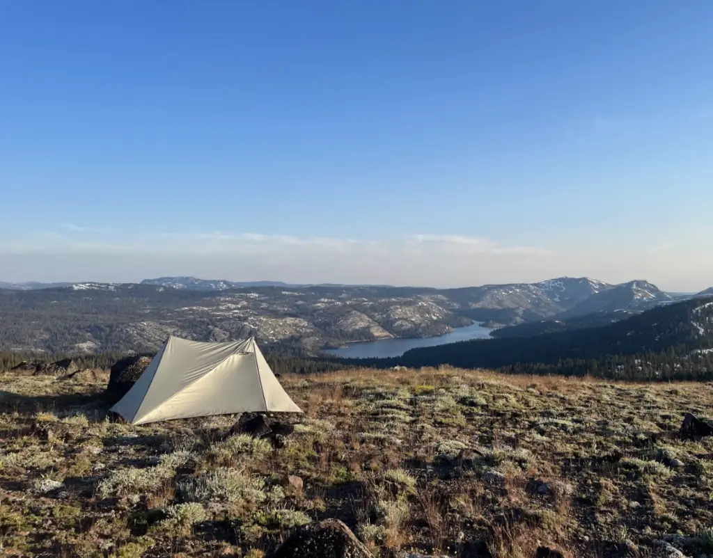 A canvas tent on a grassy hill overlooking a scenic lake and forested mountains during sunset.