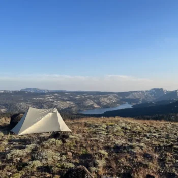 A canvas tent on a grassy hill overlooking a scenic lake and forested mountains during sunset.